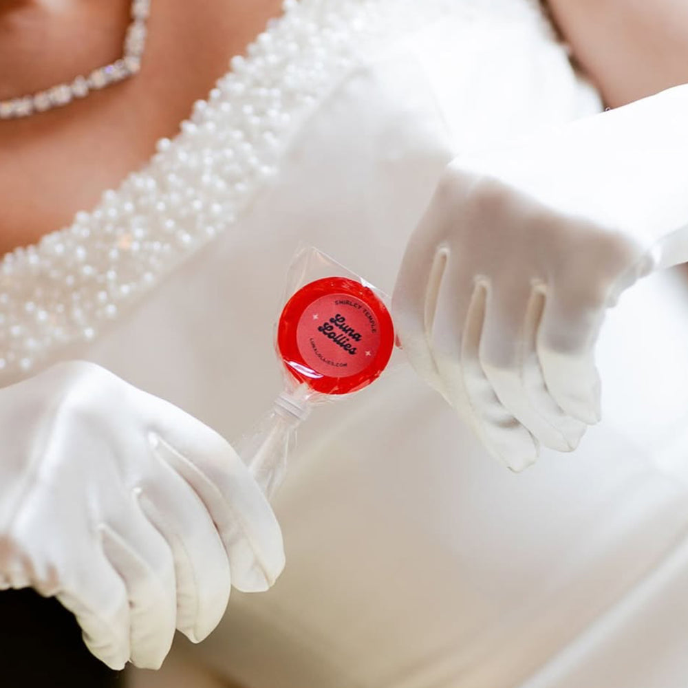 Bride wearing an elegant wedding gown and white formal gloves holding a red lollipop