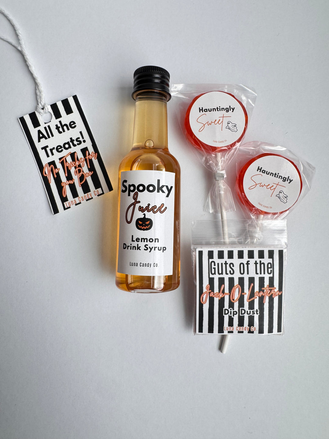 A Halloween-themed treat bundle featuring a miniature bottle of lemon drink syrup, two individually wrapped lollipops, a small bag of dip dust, and two black-and-white striped gift tags on a light gray background.