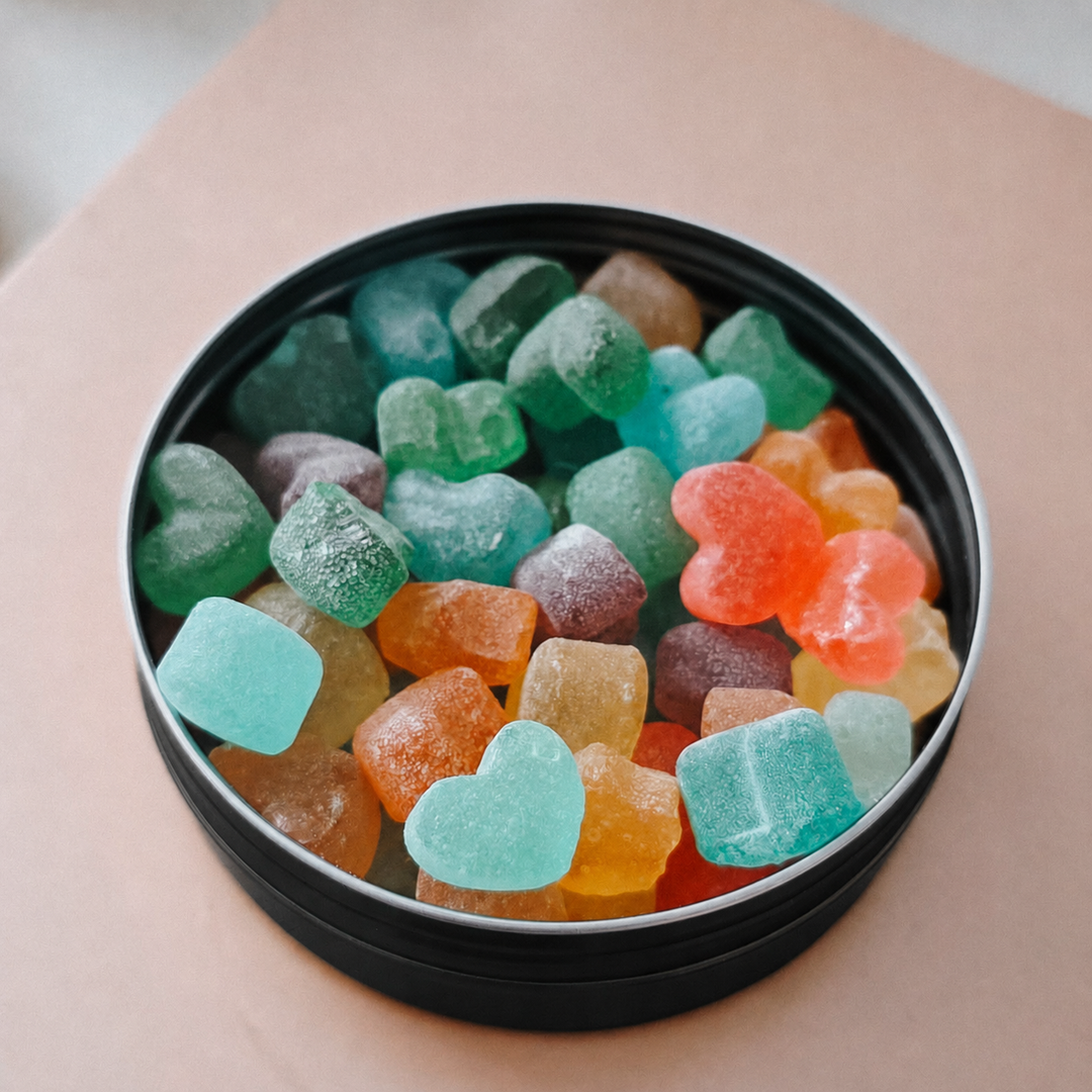 A round black tin filled with assorted heart-shaped and rounded hard candies in multiple colors, including mint green, orange, purple, and yellow, set on a peach-colored surface.