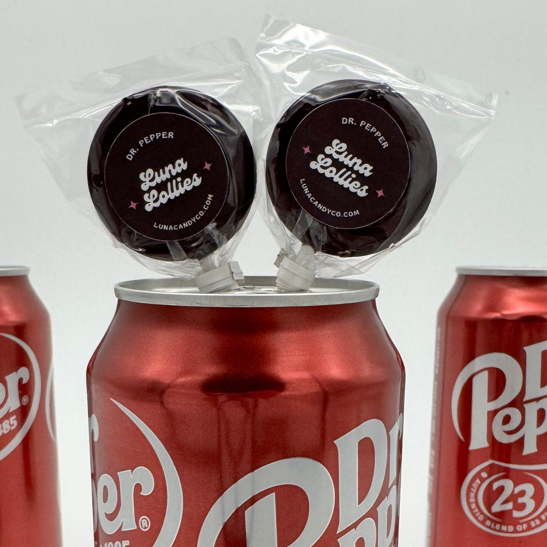 Three red Dr Pepper 23 cans on a white background with two Dr. Pepper Lollipops from Luna Candy Co.