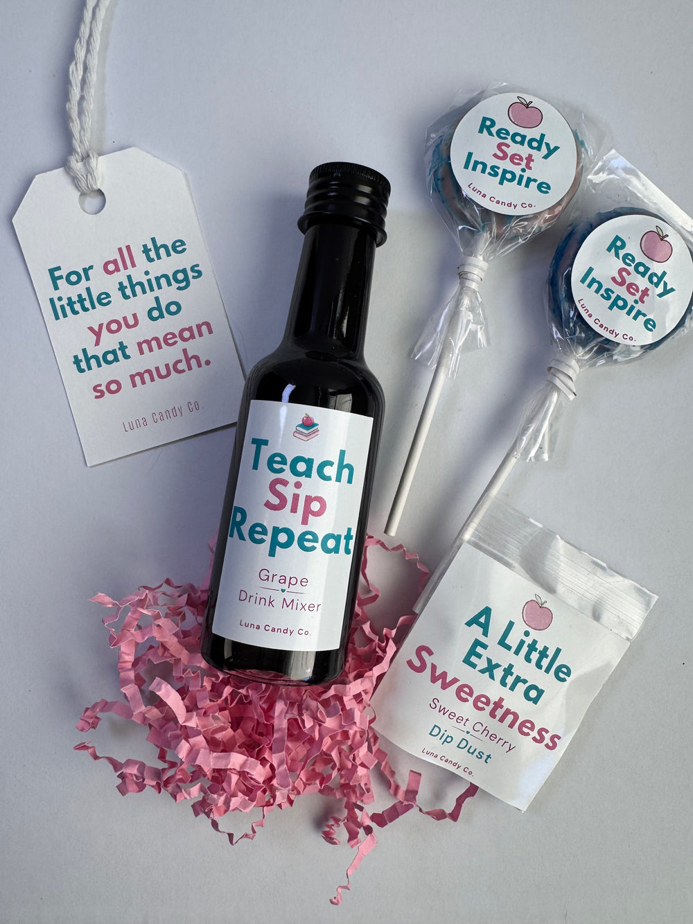 A curated gift set on a light gray background featuring a small black bottle labeled 'Teach Sip Repeat' Grape Drink Mixer, a bag of 'A Little Extra Sweetness' Dip Dust, two wrapped lollipops labeled 'Ready Set Inspire', and a hanging gift tag with the quote 'For all the little things you do that mean so much.' Pink crinkle paper is scattered around the bottle.
