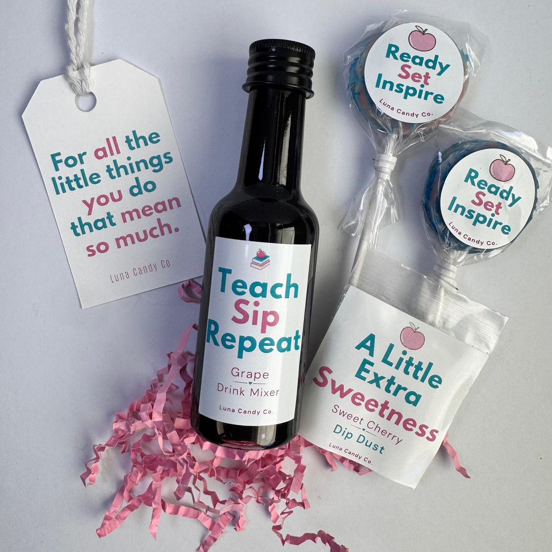 A gift set on a light gray background featuring a small dark bottle with grape drink syrup labeled 'Teach Sip Repeat Grape Drink Mixer,' two wrapped lollipops with round labels reading 'Ready Set Inspire,' a bag labeled 'A Little Extra Sweetness Dip Dust,' and a hanging tag with the phrase 'For all the little things you do that mean so much.' Pink crinkle paper is scattered beneath the items.