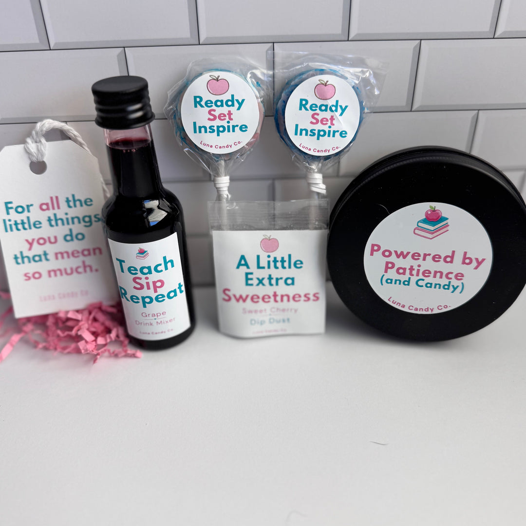A curated gift set on a white surface against a white subway tile wall, featuring a small bottle of red liquid labeled &
