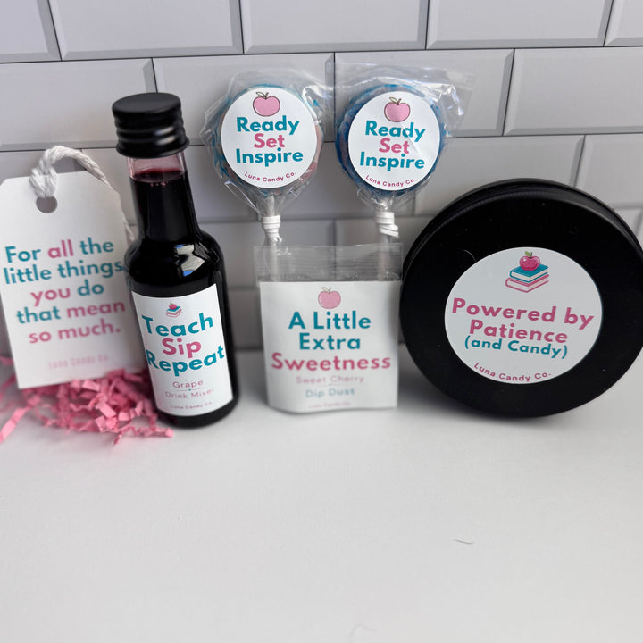 A curated gift set on a white surface against a white subway tile wall, featuring a small bottle of red liquid labeled &