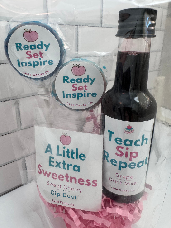 A gift set from Luna Candy Co. containing a bottle of Grape Drink Mixer, a Sweet Cherry Dip Dust packet, and two lollipops, all wrapped in clear cellophane with pink paper filler, set against a white tiled background.