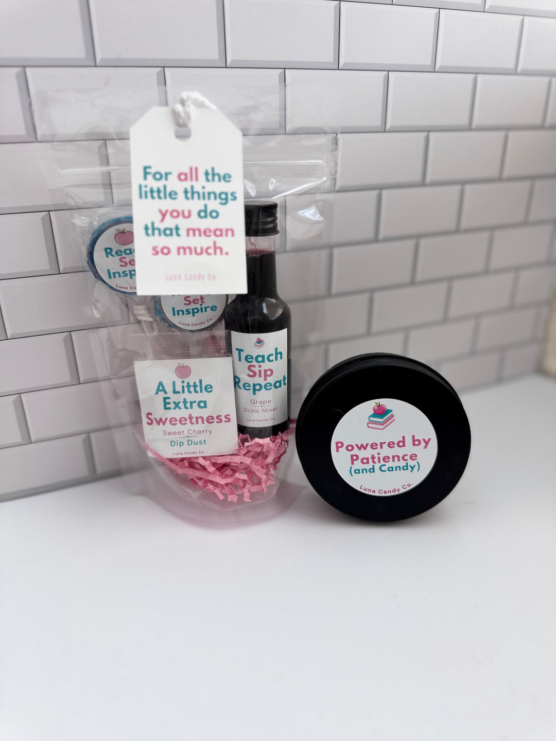 A small gift set in clear cellophane packaging on a white surface against a white subway tile wall. The set includes a black bottle labeled Teach Sip Repeat, a round black tin labeled Powered by Patience and Candy, pink paper shred, and a hanging white tag with a quote.