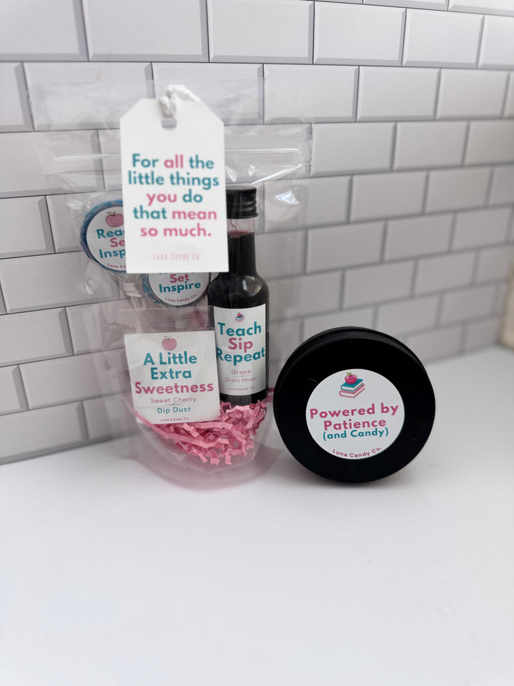 A small gift set in clear cellophane packaging on a white surface against a white subway tile wall. The set includes a black bottle labeled Teach Sip Repeat, a round black tin labeled Powered by Patience and Candy, pink paper shred, and a hanging white tag with a quote.
