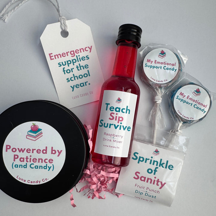 A curated gift set on a light gray background featuring a small bottle of raspberry drink mixer, a black tin of fruit dip dust, two lollipops, and paper tags with playful school-themed messages. The brand &