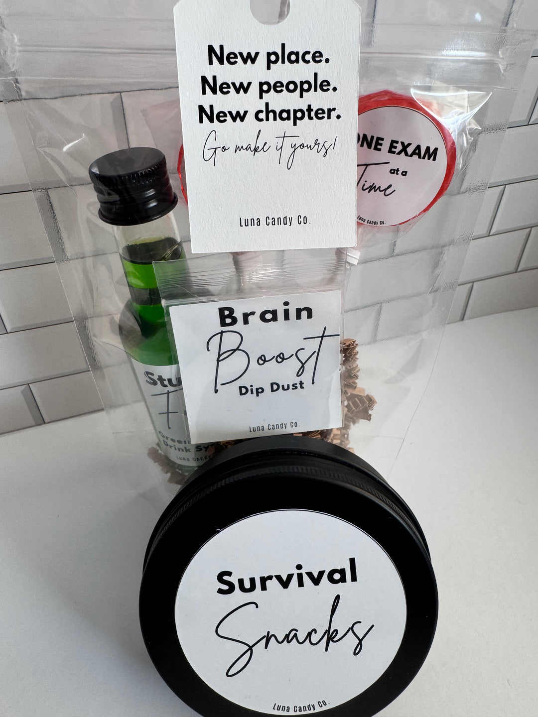 A curated snack and treat gift set in a clear plastic bag on a white countertop. The set includes a small green bottle with a black cap, a round black tin labeled &