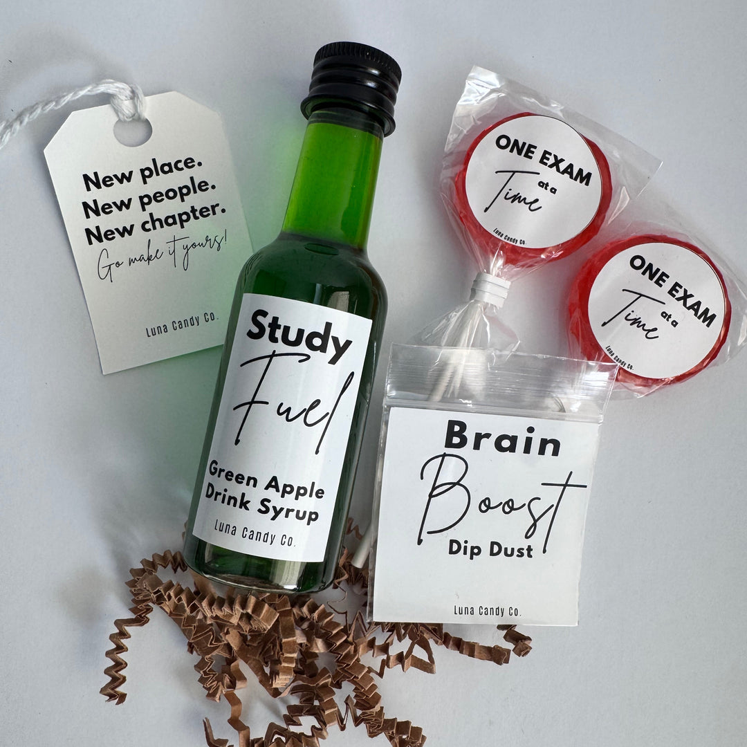 A gift set on a light gray background featuring a miniature green bottle of 'Study Fuel' green apple drink syrup, a bag of 'Brain Boost' dip dust, two red-and-white 'ONE EXAM TIME!' lollipops, and a white hanging tag with motivational text. Brown crinkle paper is scattered at the base.