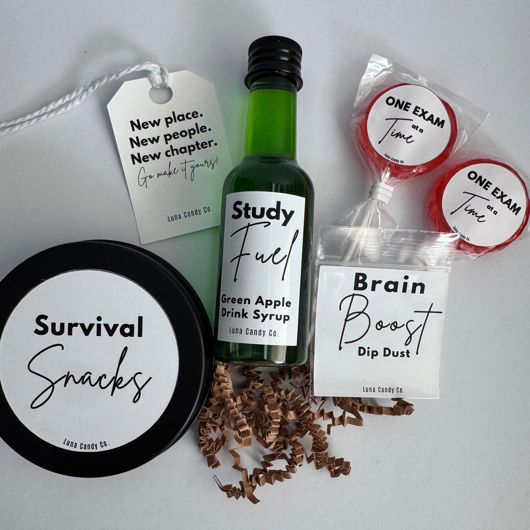 A curated snack and study-themed gift set on a light gray background, including a small green bottle labeled Study Fuel Green Apple Pink Syrup, a black round tin labeled Survival Snacks, two wrapped red lollipops labeled One Exam Twice, a clear bag labeled Brain Boost Dip Dust, and a hanging tag with a quote.