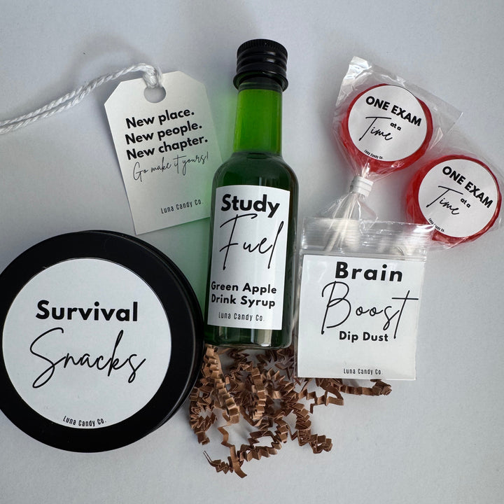 A curated gift set from Long Candy Co. for college students arranged on a light gray background, featuring a green bottle of green apple drink syrup, a black-lidded jar of survival snacks,  two brain boost dip dust packets, two lollipops, and a shredded brown filler strip.