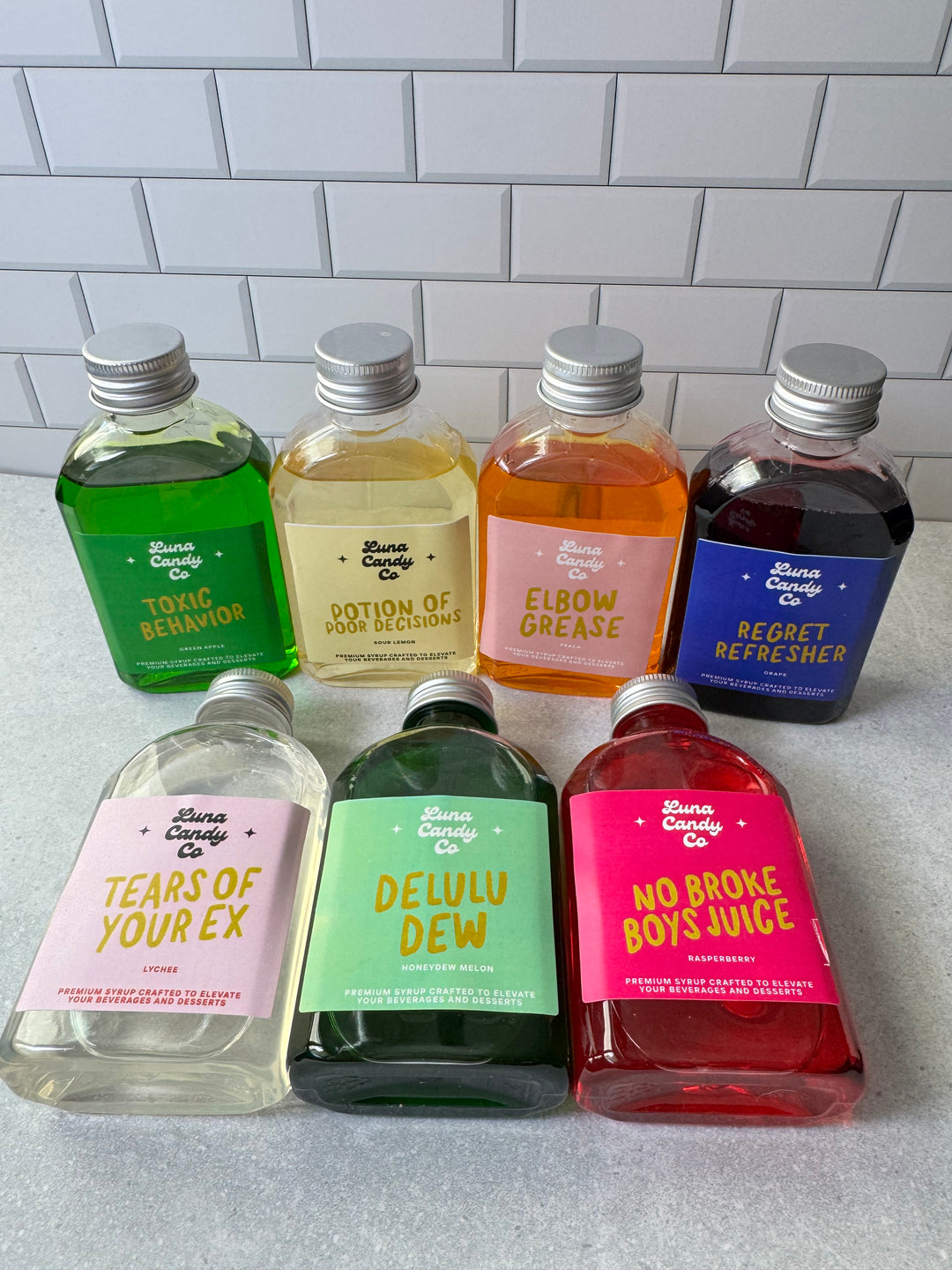 Assorted small bottles of Luna Candy Co drink syrup with colorful labels on a gray surface against a tiled wall background.